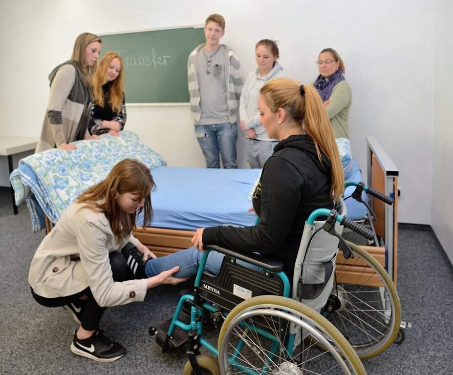 Pflegeübungen von Schülerin an einer im Rollstuhl sitzenden Person. Im Hintergrund weitere Schüler/innen hinter einem Pflegebett.